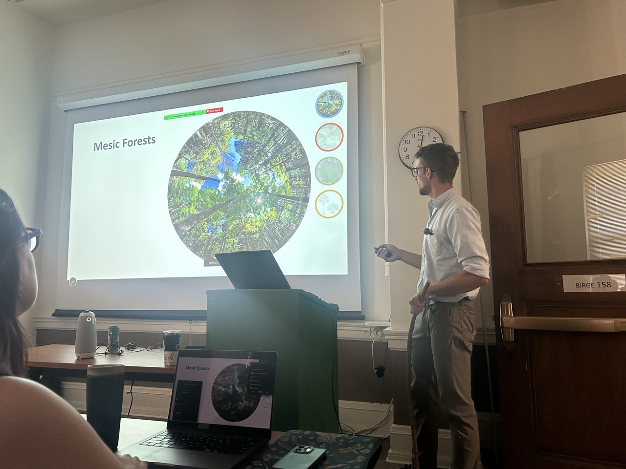 Sam Anderson stands at the front of the room facing a projector screen that displays an image of mesic forests