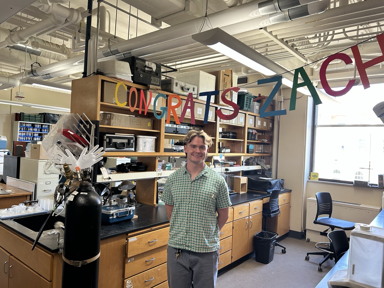 Zach stands in the lab under a "Congrats Zach" sign.