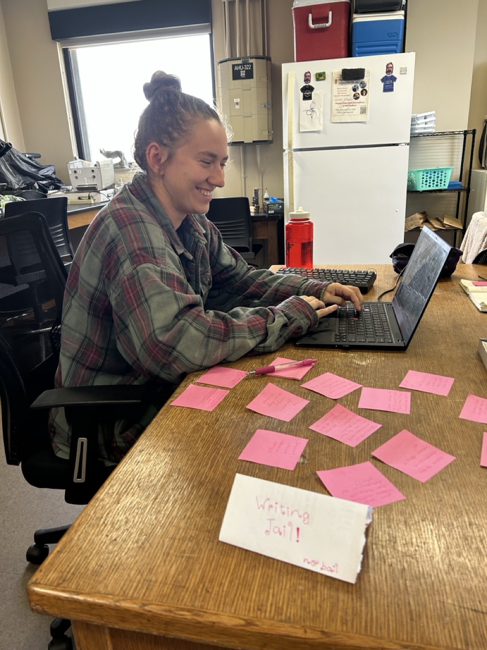 Cecilia sits at a table with a laptop computer. The table is littered with post-it notes and there is a small sign that says "Writing jail! No bail"