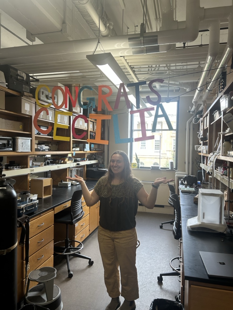 Cecilia stands in a research lab under a paper cut-out banner reading "Congrats Cecilia"
